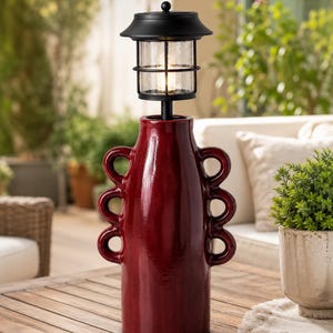 May include: A deep red ceramic vase with three circular handles on each side, topped with a black metal solar-powered lantern featuring a clear glass shade and a grid pattern. The lamp emits a warm light from the center of the glass enclosure.