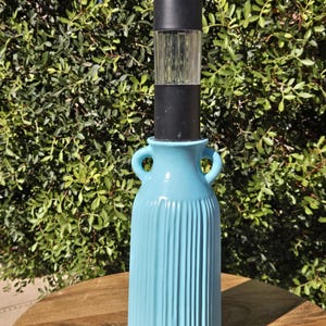 May include: A turquoise blue vase with vertical ribbed detailing, featuring two small handles and a black solar light fixture on top. The vase is set on a wooden table against a backdrop of green foliage.