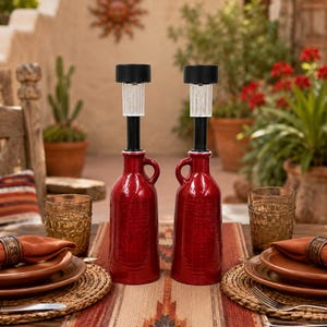 May include: Two red ceramic bottles with handles, each topped with a black solar-powered light fixture featuring a clear, vertically ribbed glass casing. The bottles are placed upright on a table, serving as decorative outdoor lighting pieces.