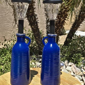 May include: Two cobalt blue ceramic bottles with integrated solar-powered lights. Each bottle features a side handle, a cylindrical body with horizontal textured ridges, and a clear, faceted light fixture mounted on a black stem at the top.