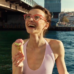 May include: A person wearing pink-framed glasses and a pink halter top smiles while holding an ice cream cone with pink and green scoops. The background includes a body of water and a bridge.