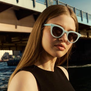 May include: A woman wearing light blue cat-eye sunglasses with dark lenses. The sunglasses have a matte finish and are set against a blurred background of a bridge and water.
