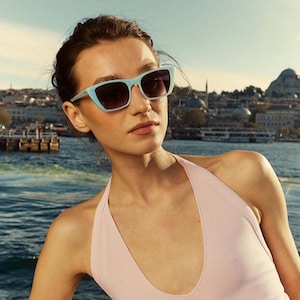Puede incluir: Una mujer con gafas de sol celeste con lentes oscuros y un top halter rosa. El fondo muestra un cuerpo de agua y un paisaje urbano bajo un cielo brillante. Las gafas de sol son rectangulares.