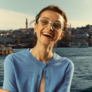 Puede incluir: Una mujer con gafas azul claro y una rebeca azul claro sonríe ampliamente. El fondo presenta un paisaje urbano con una masa de agua y un cielo despejado.