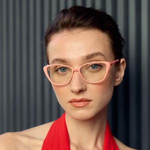 Puede incluir: Una persona con gafas rosas de ojo de gato con lentes transparentes. Las gafas tienen una montura fina y una forma ligeramente curvada hacia arriba. La persona tiene la piel clara, ojos azules y cabello oscuro. El fondo es un patrón de rayas verticales gris oscuro.