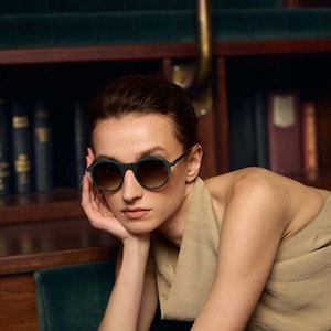 May include: A person wearing round sunglasses with dark green frames and gradient brown lenses. The person is resting their head on their hand. The background includes a dark green velvet chair and a wooden bookshelf.
