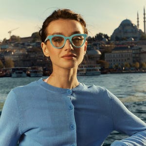 May include: A woman wearing a light blue cardigan and turquoise-framed eyeglasses. The eyeglasses have a cat-eye shape. The background features a body of water and buildings.