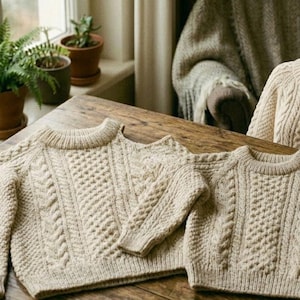 May include: Three beige cable-knit sweaters are displayed on a wooden table. The sweaters have a round neckline and a textured pattern. A cup of tea and potted plants are in the background. A mannequin is wearing a similar sweater.