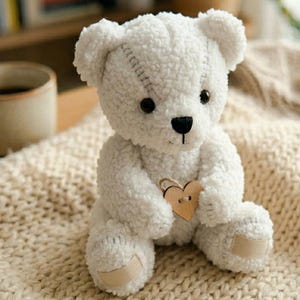 May include: A white teddy bear with a stitched design on its head and a wooden heart held in its paws. The plush toy has black eyes and a black nose. It is sitting on a cream-coloured knitted blanket.
