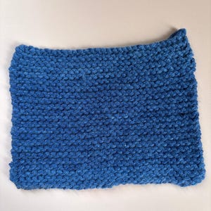 May include: A rectangular, hand-knitted item in a vibrant shade of blue. The texture is clearly visible, showcasing the individual stitches and the soft, plush quality of the yarn. The item appears to be a small blanket or washcloth.