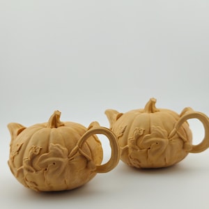 May include: Two wooden teapots shaped like pumpkins. Each teapot has a handle, a spout, and a stem. The pumpkins are light brown with carved leaf and vine details. The teapots are set against a white background.