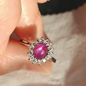 May include: A silver ring with a central oval, purplish-red gemstone, encircled by a halo of small, clear gemstones. The ring has a thin, polished band. The setting is detailed, forming a starburst around the main stone, creating a striking visual.