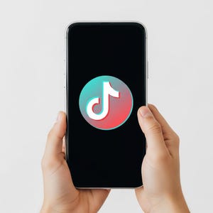 May include: A smartphone displaying the TikTok logo. The logo features a white musical note on a gradient background of red, pink, and teal. The phone is held in two hands against a white backdrop.