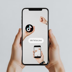May include: A smartphone displaying a TikTok logo and the text "500 TikTok Likes." The phone is held in two hands, with a smaller image of the phone on the screen. The background is a plain, light gray.