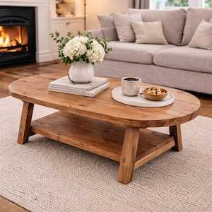 May include: A rustic, oval-shaped wooden coffee table with a lower shelf. The table holds a white vase with white flowers, books, a tray with a cup, and a small bowl of nuts. The table sits on a light-colored rug.