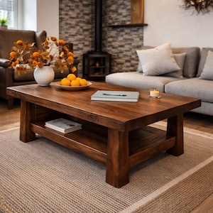 Puede incluir: Una mesa de centro de madera marrón oscuro con un estante inferior. La mesa rectangular tiene patas gruesas. Un jarrón con flores naranjas, un cuenco de naranjas y libros están sobre la mesa. La mesa está sobre una alfombra beige. La mesa mide unos 120 cm de largo.
