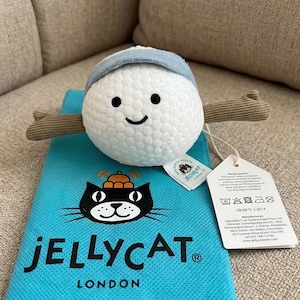 May include: A plush toy with a white ball-shaped body, a blue hat, and brown stick-like arms. It has a smiling face with black eyes. The toy rests on a turquoise bag with the "JELLYCAT LONDON" logo and a black cat graphic.