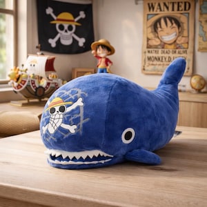 May include: A plush blue whale toy with a white and yellow pirate flag design on its side. The whale has a white mouth with teeth and a single black eye. The background includes a pirate flag and a "Wanted" poster.