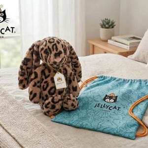 May include: A soft toy leopard-print bunny with long ears and a pink nose. It has a tag attached. Next to it is a light blue drawstring bag with a cat logo and the text "Jellycat London". The items are on a bed.