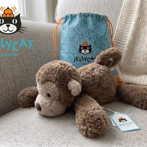 May include: A brown plush monkey toy with a light brown face and ears, lying on a gray sofa. A blue drawstring bag with a cartoon pattern and the Jellycat logo is behind the monkey. A Jellycat tag is attached.
