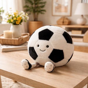 May include: A plush soccer ball with a smiling face, black and white panels, and small, dotted feet. The soft toy is designed to sit upright and is made of a soft, fuzzy material. A cute and cuddly children's toy.