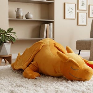 Könnte beinhalten: Ein großes, plüschiges, orangefarbenes Charizard-Kissen liegt auf einem weißen Shag-Teppich. Das Kissen hat Flügel, einen Schwanz und ein lächelndes Gesicht. Der Raum hat eine neutrale Farbpalette mit einer Pflanze und gerahmten Kunstwerken.