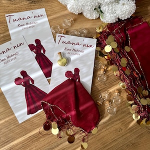 Puede incluir: Una colección de recuerdos y decoraciones de boda sobre una superficie de madera. La imagen incluye tarjetas de invitación con la silueta de una mujer con un vestido rojo, una bufanda roja con lentejuelas doradas, pequeñas botellas de vidrio y flores blancas.