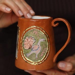 Puede incluir: Una taza de cerámica con exterior marrón e interior blanco. La taza presenta un diseño pintado a mano de un pájaro con cola rosa y fondo verde, enmarcado con ribete dorado. El asa también es marrón.