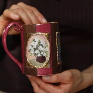 Peut inclure: Une tasse en céramique décorative avec une peinture florale. La tasse a une anse bordeaux et des panneaux alternés marron et bordeaux. La peinture représente des fleurs blanches dans un vase marron, encadrées d'accents dorés.