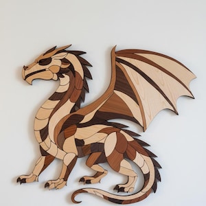 Może przedstawiać: Drewniana grafika ścienna przedstawiająca smoka w odcieniach brązu, beżu i ciemnego drewna. Smok ma szczegółowe skrzydła, kolczysty grzbiet i jest wykonany z różnych kawałków drewna. Grafika jest zamontowana na białym tle.
