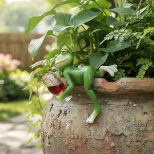 Puede incluir: Una caprichosa figura de rana verde con patas blancas, sosteniendo una copa de vino tinto, posada en el borde de una maceta de terracota llena de exuberantes plantas verdes. La pose relajada de la rana añade un toque de humor a la decoración del jardín.