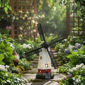 Puede incluir: Un molino de viento decorativo para jardín con cuerpo blanco, base roja y aspas negras. El molino tiene pequeñas ventanas iluminadas. Se encuentra en un camino de piedra rodeado de vegetación y flores, creando una escena exterior encantadora.