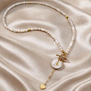 May include: White beaded necklace with gold accents and a circular pendant featuring the letter "A". The necklace has a toggle clasp and a small gold heart charm. The jewelry is displayed on a satin surface.