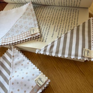 May include: Three triangular fabric cloths with pinked edges, featuring beige and white patterns including stripes, polka dots, and small flowers. Each cloth has a small beige fabric tag sewn on the edge with the text Nini's Corner printed in black.
