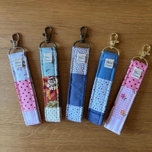 May include: Five fabric wristlet keychains with metal lobster clasps. Each strap features a patchwork design of various patterns including polka dots, floral prints, and stripes in pink, blue, and brown tones. Each has a small label reading Nini's Corner.