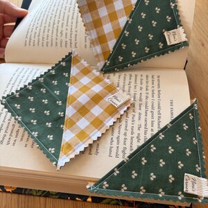 May include: Three triangular fabric corner bookmarks placed on an open book. Each bookmark features a combination of dark green fabric with a small white floral pattern and yellow and white gingham check fabric, finished with pinked edges and a small tag.