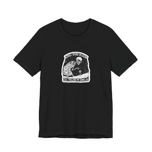 Könnte beinhalten: Schwarzes T-Shirt mit einem weißen Aufdruck eines Skeletts, das in eine Decke gehüllt nach einer Blume greift. Der Text "ALMOST SPRING DELUSION" steht über dem Bild, darunter "STILL FREEZING MY BONES OFF".