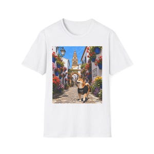 May include: A white t-shirt featuring a centered graphic print of a Shiba Inu dog wearing a black vest, standing on a cobblestone street in a narrow alleyway lined with white walls, blue flower pots, and vibrant red flowers under a blue sky.