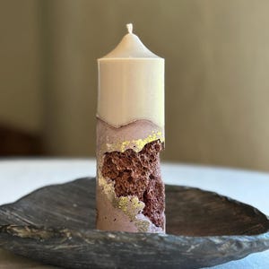 Peut inclure: Une bougie cylindrique composée d'une partie supérieure lisse de couleur crème et d'une base texturée mauve. Une découpe irrégulière et poreuse révèle un intérieur brun foncé, avec des bords rehaussés de feuilles d'or métallisées.