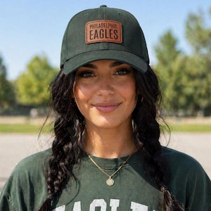 May include: Dark green baseball cap with a brown leather patch featuring the text "PHILADELPHIA EAGLES". The cap is worn by a person with dark hair and a green shirt.
