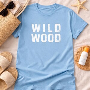 K&ouml;nnte beinhalten: Hellblaues T-Shirt mit dem wei&szlig;en Aufdruck "WILD WOOD". Das Bild zeigt Strandaccessoires: eine Strohtasche, Sonnenbrille, Sandalen und Sonnencreme. Ein Seestern und ein Palmblatt erg&auml;nzen das Strandthema und vermitteln Urlaubsstimmung.