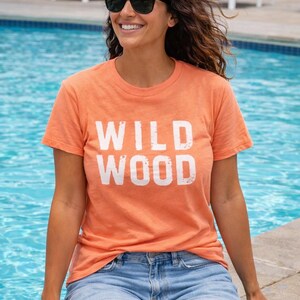 K&ouml;nnte beinhalten: Ein orangefarbenes T-Shirt mit dem wei&szlig;en Schriftzug "WILD WOOD". Die Person tr&auml;gt blaue Jeansshorts und eine schwarze Sonnenbrille. Im Hintergrund ein Swimmingpool und Liegest&uuml;hle.
