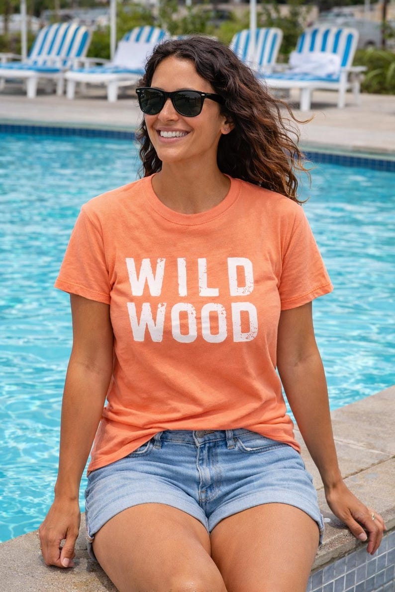 K&ouml;nnte beinhalten: Ein orangefarbenes T-Shirt mit dem wei&szlig;en Schriftzug "WILD WOOD". Die Person tr&auml;gt blaue Jeansshorts und eine schwarze Sonnenbrille. Im Hintergrund ein Swimmingpool und Liegest&uuml;hle.