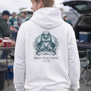 Könnte beinhalten: Weißer Kapuzenpullover mit einem Grafikdesign auf der Rückseite, das einen Adler, ein Fernglas und den Text "PHILADELPHIA BIRD WATCHING CLUB" zeigt. Das Design ist in Türkis- und Grautönen gehalten. Der Pullover wird im Freien getragen.