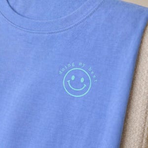 May include: A light blue t-shirt with a crew neck. The shirt features a teal smiley face graphic with the words "doing my best" in a circular design around the smiley face.