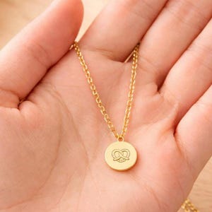 Op de afbeelding: Goudkleurige ketting met een ronde hanger met een pretzel-ontwerp. De hanger hangt aan een delicate ketting. De ketting wordt in een hand gehouden, met een vage achtergrond.