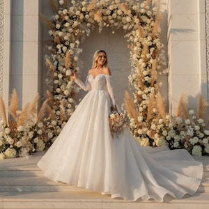 Op de afbeelding: Een witte trouwjurk met een off-the-shoulder halslijn en lange mouwen. De jurk heeft een volle rok en een lange sleep. Het lijfje en de mouwen zijn versierd met kant. De bruid houdt een boeket vast voor een bloemenboog.