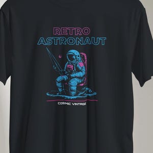 Op de afbeelding: Zwart t-shirt met een retro astronaut ontwerp. De afbeelding toont een visserij astronaut in roze en blauw, met de tekst "RETRO ASTRONAUT" en "COSMIC VINTAGE".