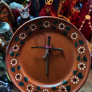 May include: A round terracotta plate featuring a hand-painted border of white flowers and dark green leaves. Centered on the plate is a cross constructed from two dark, weathered metal nails bound together with a thin, dark pink cord.