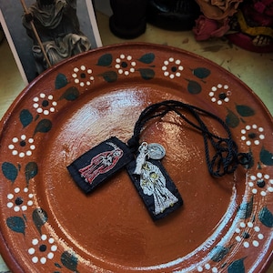May include: A black cord necklace featuring two rectangular fabric pendants with embroidered figures of a robed entity, one in red and one in white, accompanied by two small silver-toned metal charms, one shaped like a figure and one circular.